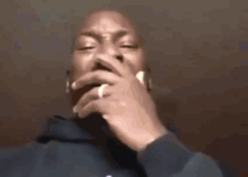A Man In A Blue Hoodie Is Smoking A Cigarette With His Hand On His Mouth . GIF