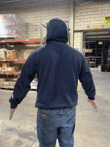 A Man In A Blue Hoodie Is Standing In A Warehouse With His Arms Outstretched In Front Of A Sign That Says Exit GIF