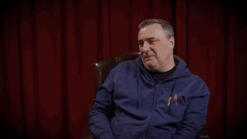 A Man In A Blue Hoodie Sits In Front Of A Red Curtain With The Words " Wide Beam That On Kil " Behind Him GIF