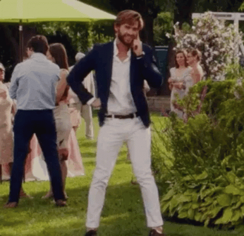A Man In A Blue Jacket And White Pants Is Standing In A Grassy Field Talking On A Cell Phone . GIF