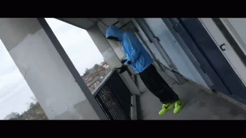 A Man In A Blue Jacket And Yellow Nike Shoes Is Standing On A Balcony GIF
