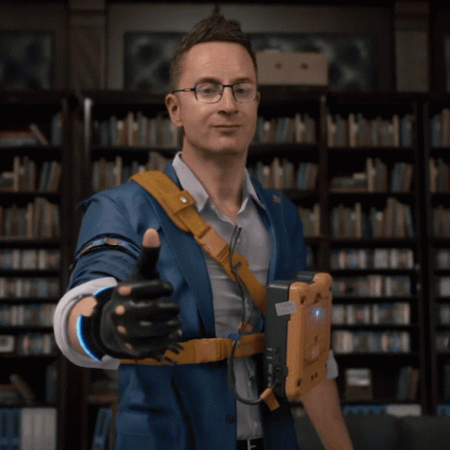 A Man In A Blue Jacket Giving A Thumbs Up In Front Of A Bookshelf GIF