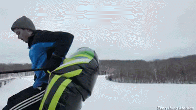 A Man In A Blue Jacket Is Pulling Another Man On A Sled GIF