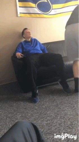 A Man In A Blue Jacket Is Sitting On A Black Couch In Front Of A Banner That Says Tm GIF