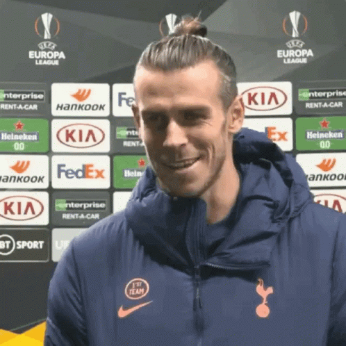 A Man In A Blue Jacket Is Smiling In Front Of A Europa League Banner GIF