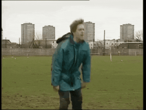 A Man In A Blue Jacket Is Standing In A Field With Tall Buildings In The Background GIF