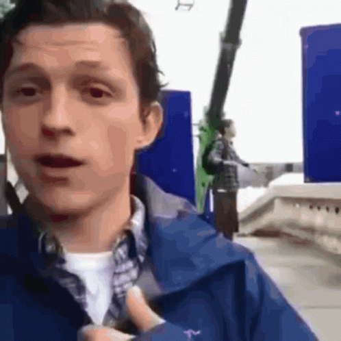 A Man In A Blue Jacket Is Standing In Front Of A Roller Coaster And Pointing At It . GIF