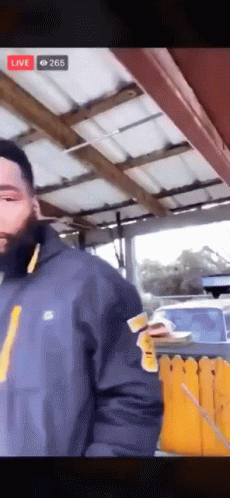 A Man In A Blue Jacket Is Standing In Front Of A Yellow Fence While Holding A Piece Of Paper . GIF