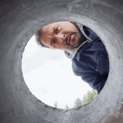 A Man In A Blue Jacket Looks Through A Hole In A Pipe GIF