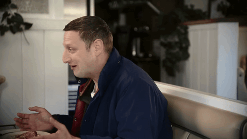 A Man In A Blue Jacket Sits At A Table With His Hands Out GIF