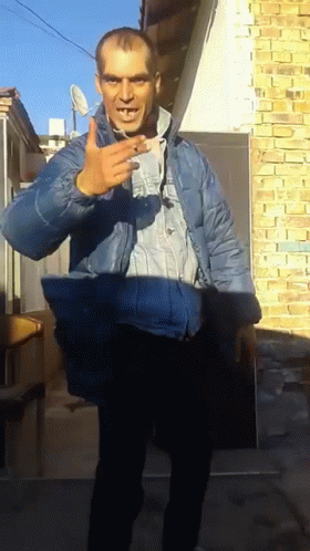 A Man In A Blue Jacket Stands In Front Of A Brick Building GIF