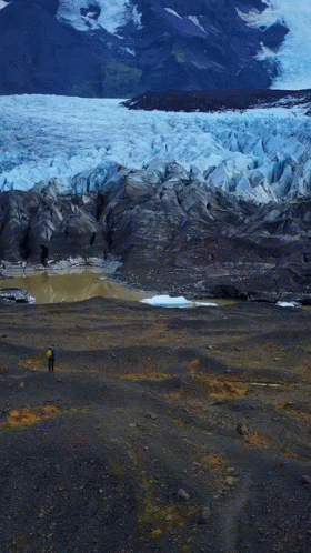 A Man In A Blue Jacket Stands In Front Of A Glacier GIF