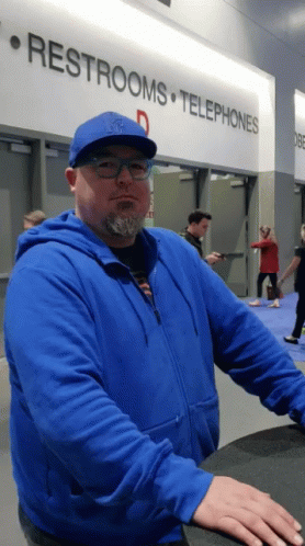A Man In A Blue Jacket Stands In Front Of A Sign That Says Restrooms And Telephones GIF