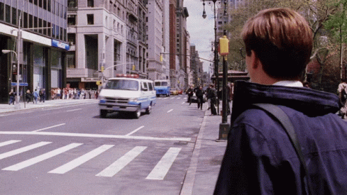 A Man In A Blue Jacket Stands On The Sidewalk Looking At A Crosswalk GIF