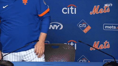 a man in a blue mets jersey sits at a table with microphones in front of a wall that says citi field