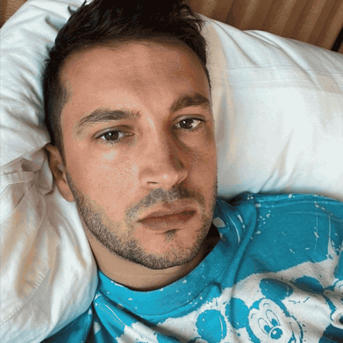 A Man In A Blue Mickey Mouse Shirt Is Laying On A Bed GIF