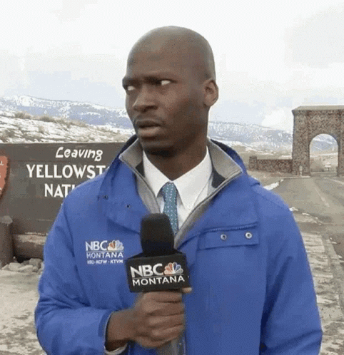 A Man In A Blue Nbc Montana Jacket Holds A Microphone In Front Of A Sign That Says Leaving Yellowstone National Park GIF