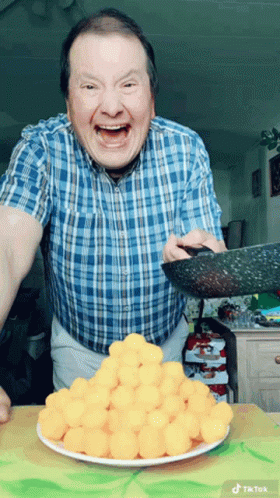 A Man In A Blue Plaid Shirt Is Holding A Pan Over A Plate Of Cheese Balls GIF
