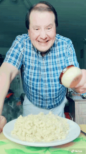 A Man In A Blue Plaid Shirt Is Pouring A Slice Of Onion On A Plate Of Macaroni And Cheese GIF