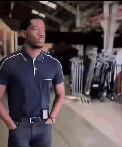 A Man In A Blue Polo Shirt Is Standing In A Warehouse With His Hands In His Pockets GIF