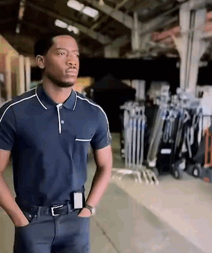 A Man In A Blue Polo Shirt Is Standing With His Hands In His Pockets In A Warehouse . GIF