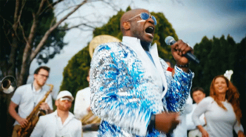 A Man In A Blue Sequined Jacket Sings Into A Microphone In Front Of A Band GIF