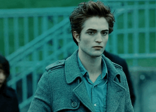 A Man In A Blue Shirt And A Grey Coat Stands In Front Of A Green Fence GIF