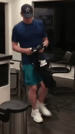A Man In A Blue Shirt And A Hat Is Standing In A Room Holding A Bottle . GIF