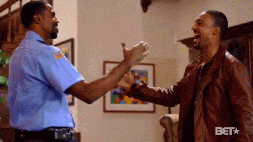 A Man In A Blue Shirt And A Man In A Brown Jacket Are Shaking Hands In A Living Room . GIF