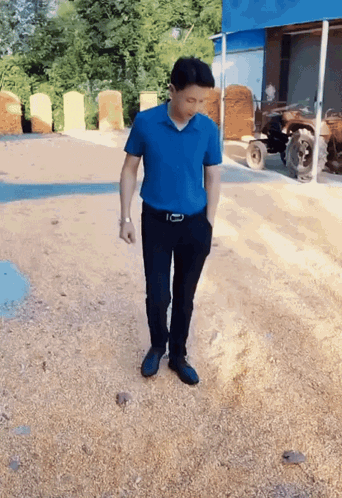 A Man In A Blue Shirt And Black Pants Is Standing On A Gravel Road GIF