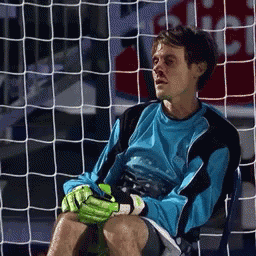 A Man In A Blue Shirt And Green Gloves Sits In Front Of A Soccer Goal GIF
