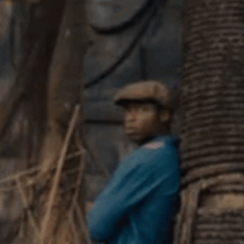 A Man In A Blue Shirt And Hat Is Standing Next To A Tree . GIF