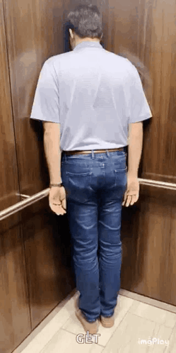A Man In A Blue Shirt And Jeans Is Standing In An Elevator . GIF