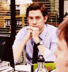 A Man In A Blue Shirt And Tie Is Sitting At A Desk With His Hand On His Chin . GIF