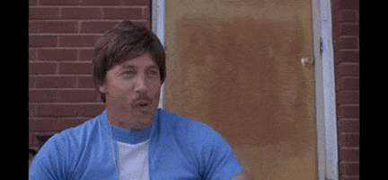 A Man In A Blue Shirt And White Shirt Is Sitting In Front Of A Brick Building . GIF