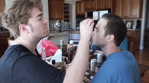 A Man In A Blue Shirt Is Applying Makeup To Another Man In A Kitchen GIF