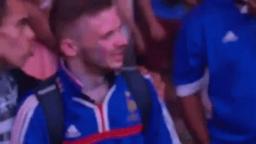 A Man In A Blue Shirt Is Covering His Face With His Hand While Standing In A Crowd . GIF