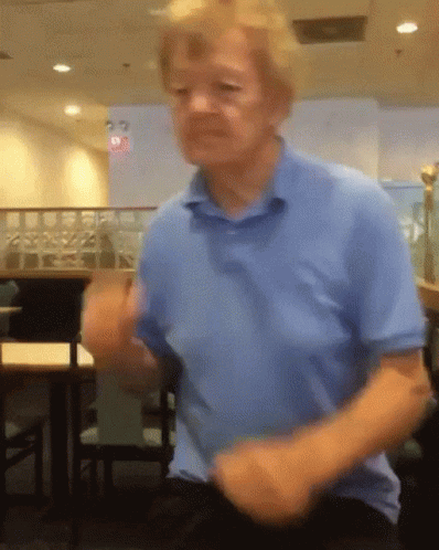 A Man In A Blue Shirt Is Dancing In A Room With Tables And Chairs GIF