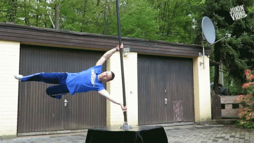 A Man In A Blue Shirt Is Doing A Handstand On A Pole In Front Of A Garage GIF