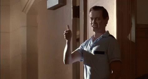 A Man In A Blue Shirt Is Giving A Thumbs Up While Standing In Front Of A Door . GIF