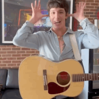 a man in a blue shirt is holding a guitar in front of a brick wall