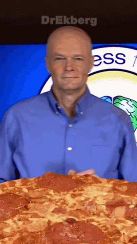 A Man In A Blue Shirt Is Holding A Large Pepperoni Pizza In Front Of A Sign That Says Ess GIF