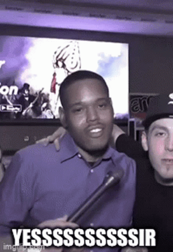 A Man In A Blue Shirt Is Holding A Microphone And Smiling While Another Man Looks On . GIF