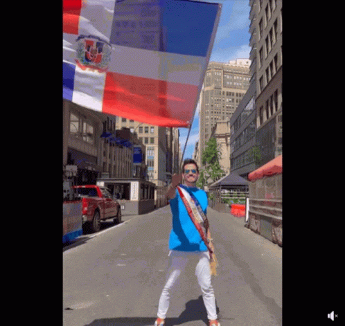 A Man In A Blue Shirt Is Holding A Red White And Blue Flag On A City Street GIF