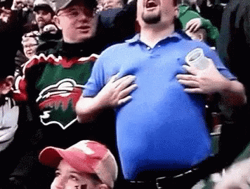A Man In A Blue Shirt Is Holding His Chest While Standing In A Crowd Of People . GIF