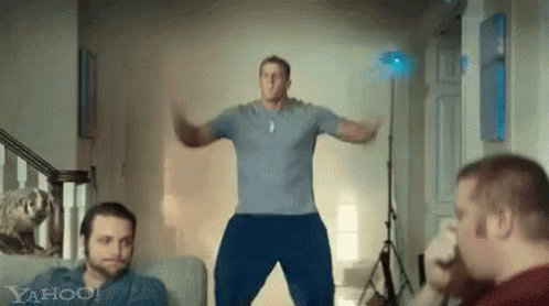 A Man In A Blue Shirt Is Jumping In The Air In A Living Room With Two Other Men . GIF