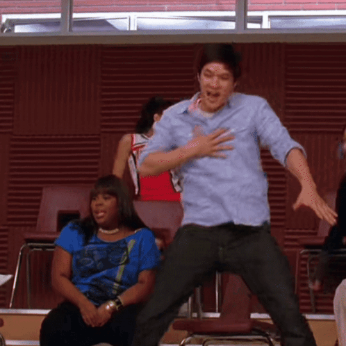 A Man In A Blue Shirt Is Jumping In The Air While A Woman Sits Behind Him GIF