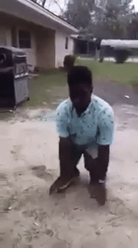 A Man In A Blue Shirt Is Kneeling Down In The Dirt In Front Of A House . GIF