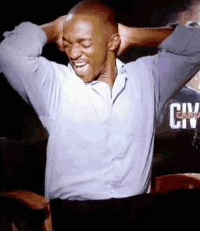 A Man In A Blue Shirt Is Laughing With His Hands Behind His Head In Front Of A Sign That Says City GIF