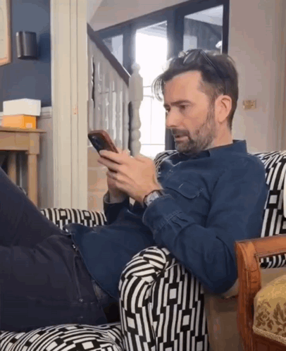A Man In A Blue Shirt Is Laying On A Chair Looking At His Phone GIF